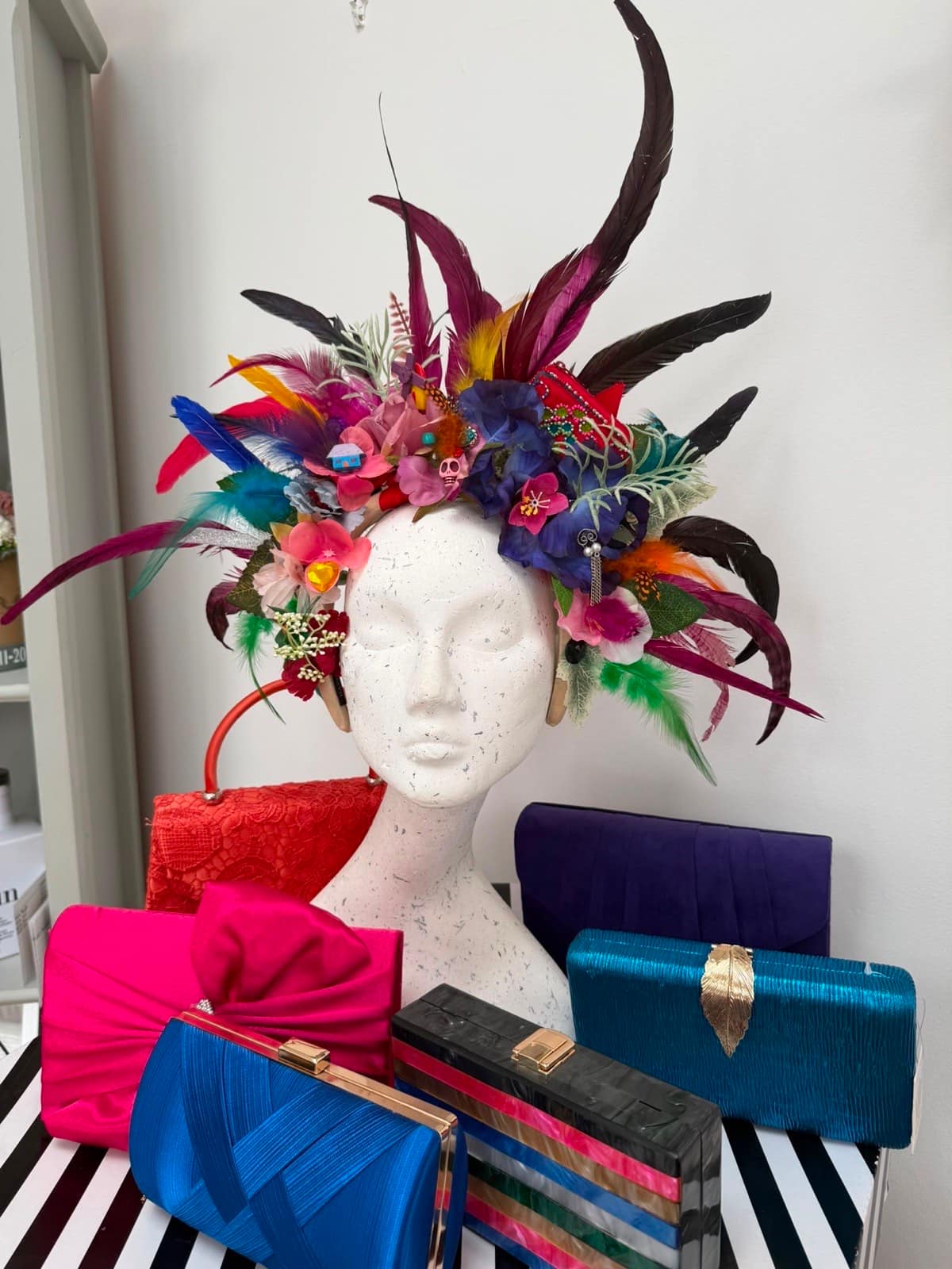Carnival-style feather headdress in magenta, blue and green with flowers and trinkets, shown with rainbow-coloured clutch bags