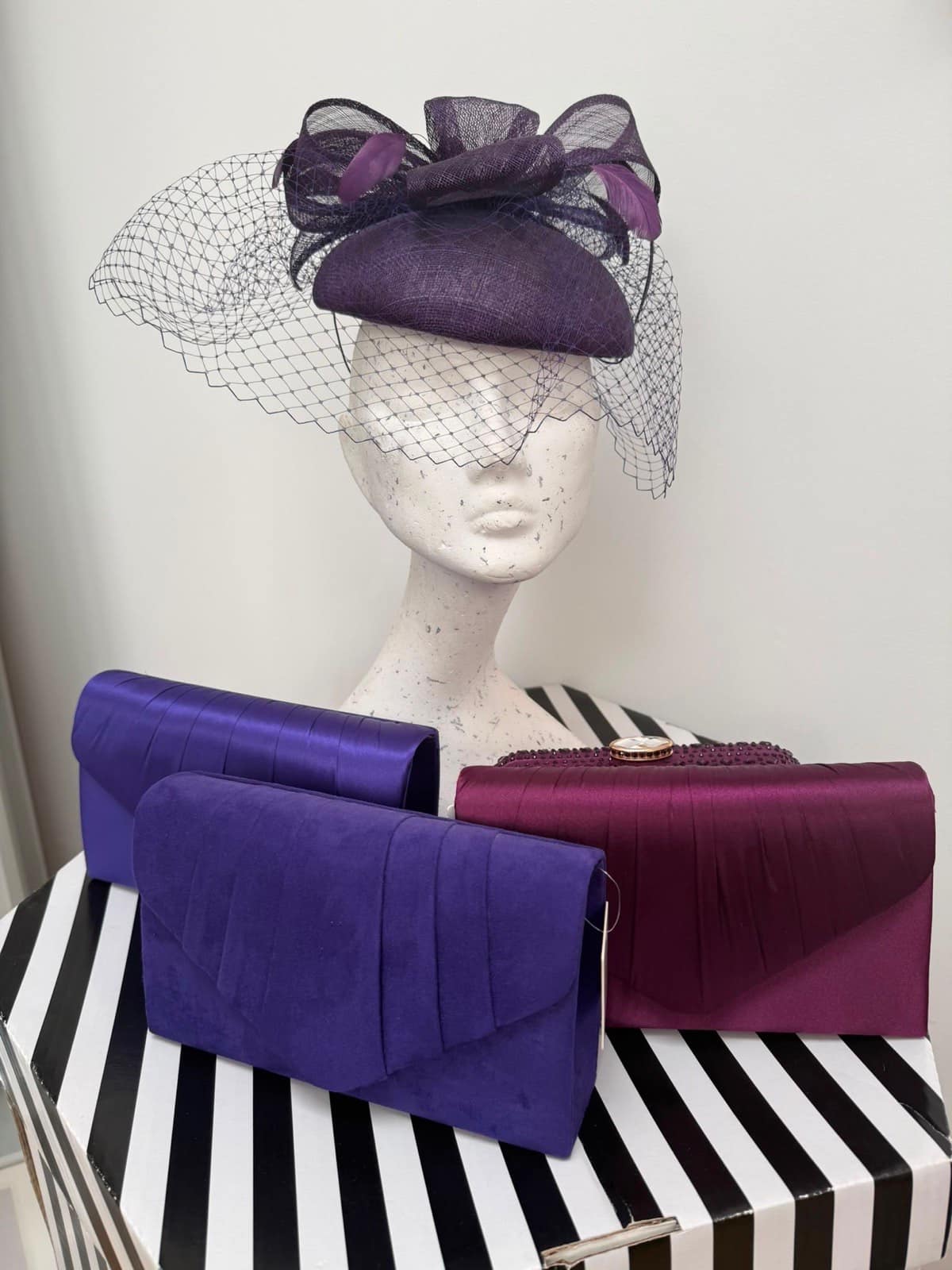 Dark plum pillbox hat with birdcage veil, bows and feathers, alongside purple and plum clutches