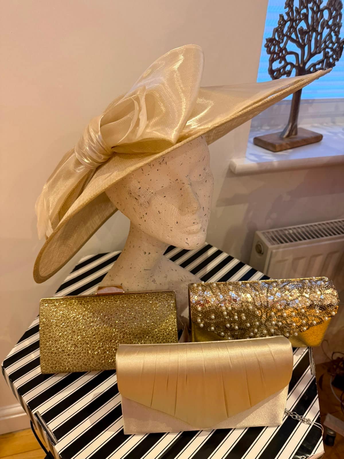 Close view of the cream organza-bow occasion hat on a mannequin with gold sparkle and pearl clutches